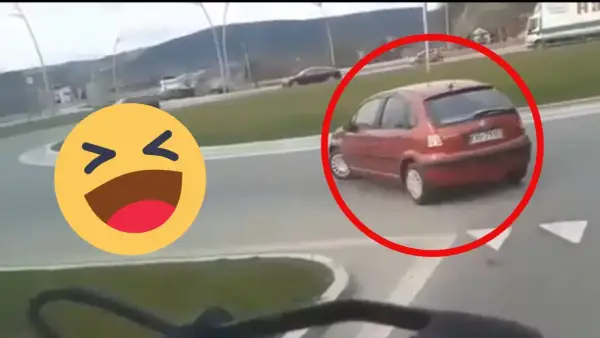 Czerwony samochód próbuje zawrócić na rondzie, ale robi to na lewo, co wygląda na niezbyt przemyślane. Obok śmieje się emotikon.