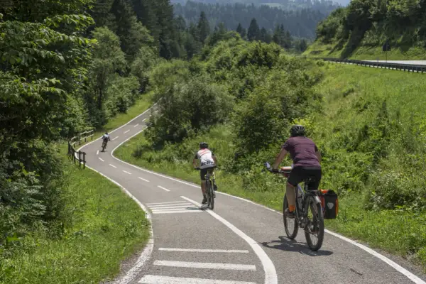 Rowerzyści przemierzają malowniczą trasę rowerową Alpe Adria, otoczeni bujną zielenią i górskim krajobrazem.