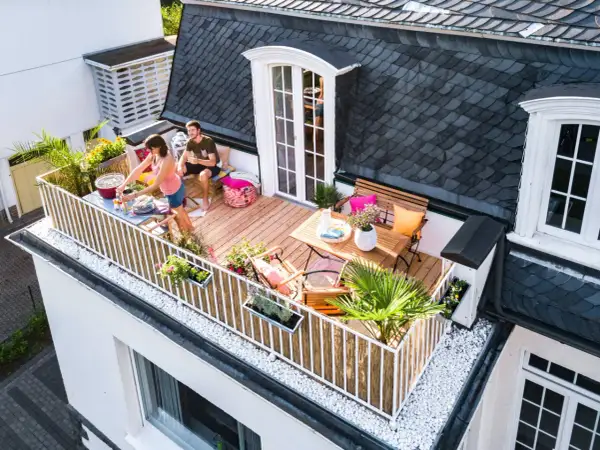 Ein Paar genießt ein Grillfest auf ihrem neu gestalteten Balkon. Der Balkon ist mit Holzmöbeln, Pflanzen und Dekorationen geschmückt.
