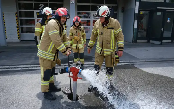 Jugendfeuerwehr übt den Umgang mit dem Hydranten. Wasser spritzt aus dem roten Standrohr, während die jungen Brandschützer in voller Montur zuschauen.