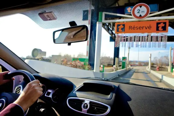 Vue depuis l'intérieur d'une voiture approchant une voie de péage. Le tableau de bord et le volant sont visibles. Un badge de télépéage est fixé au pare-brise. Le panneau "Réservé" indique comment ça fonctionne.