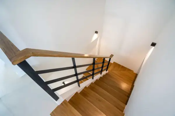 Nowoczesne schody z drewnianymi stopniami i czarną balustradą. Jak oświetlić schody w domu? Dyskretne kinkiety na ścianie tworzą nastrojowy blask.