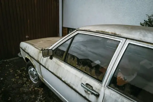 Vieille voiture blanche, couverte de poussière et de mousse, devant un garage. Un symbole de l'interdiction voiture thermique.