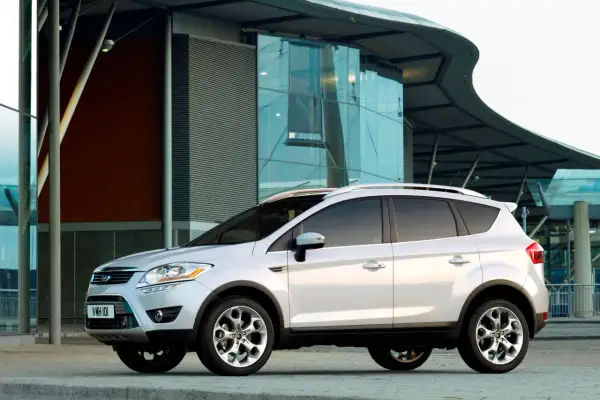 Biały Ford Kuga 2010 zaparkowany przed nowoczesnym budynkiem z przeszkloną fasadą.