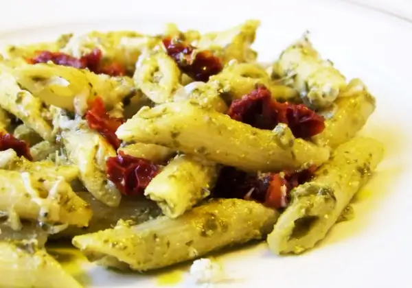 Makaron penne z zielonym pesto i suszonymi pomidorami na białym talerzu.