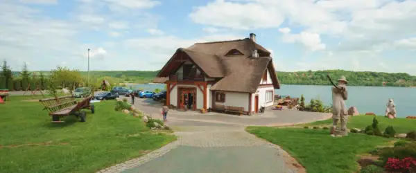 Dom z piękną, naturalną strzechą na dachu, położony nad jeziorem. Wokół zielony trawnik, drewniany wóz i rzeźby.