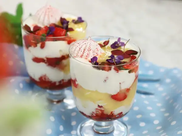 Två glas med Eton Mess, lager av vispad grädde, jordgubbar och maräng, toppade med blommor.