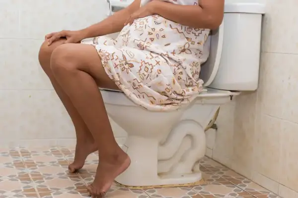 Ciężarna kobieta siedzi na toalecie, doświadczając biegunki w ciąży. Jej dłonie spoczywają na brzuchu.