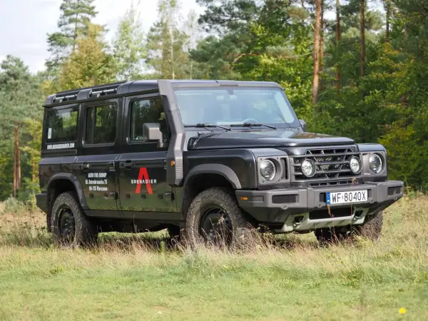 Czarny INEOS Grenadier z logo "A" na drzwiach, zaparkowany na trawie, z lasem w tle.