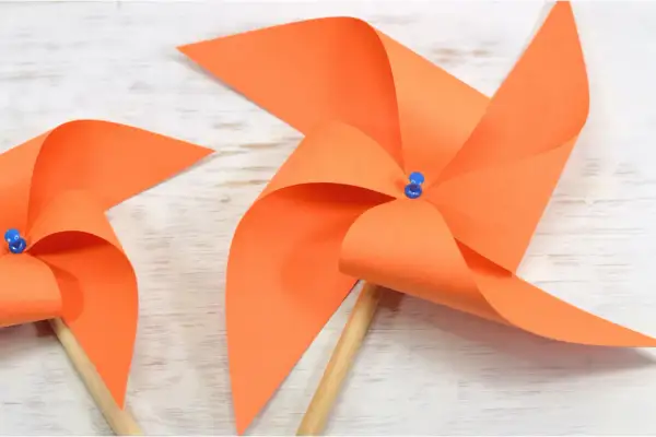 Zwei selbstgebastelte Windräder aus orangefarbenem Papier auf Holzuntergrund. Perfekt zum Windrad basteln mit Kindern.