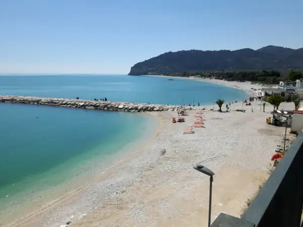 Mattinata serena sulla spiaggia di piana, con il mare azzurro che lambisce la costa ghiaiosa e il molo di sassi.