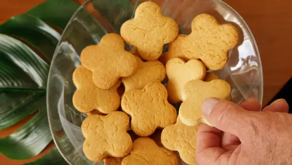 Una mano prende uno dei tanti biscotti fatti in casa a forma di quadrifoglio, disposti in una ciotola di vetro con foglie verdi sullo sfondo.