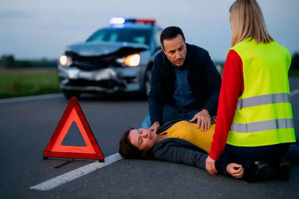 Un hombre y una mujer asisten a una persona en el suelo tras un accidente. Un triángulo de señalización y un coche dañado en el fondo ilustran la importancia de la seguridad vial.