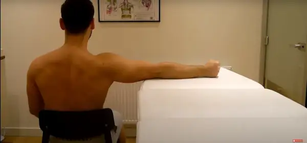 Man sitter och utför en övning för att stretcha skulderblad. Armbågen är böjd och handen vilar på en vit yta.