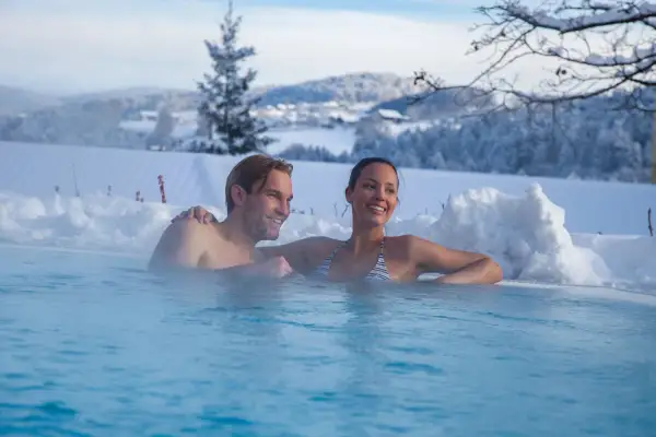 Ein Paar entspannt in einem beheizten Pool im Schnee mit verschneiter Landschaft im Hintergrund.