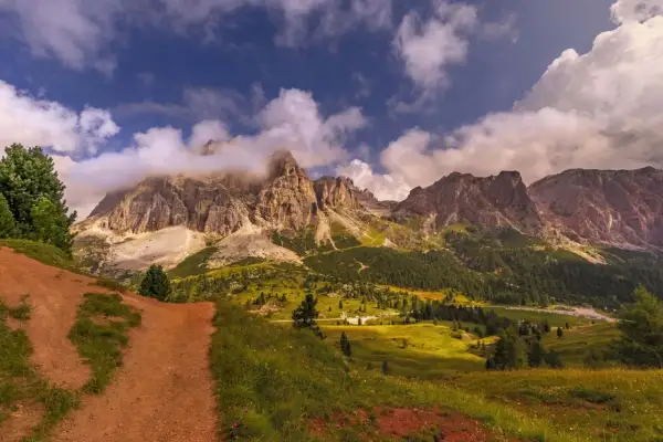 Camino de tierra rojiza que se adentra en un valle alpino con imponentes montañas cubiertas de nubes. La ruta Cinque Torri se revela entre prados verdes y bosques frondosos.