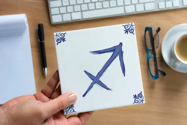 Dłoń trzyma kwadratową płytkę z symbolem Strzelca, znakiem zodiaku dla osób urodzonych 28 listopada.