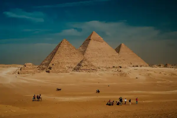 Pustynia Giza z piramidą Cheopsa w tle. Ludzie i wielbłądy przemierzają piaski pod błękitnym niebem.