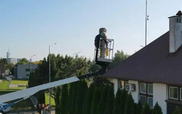 Osoba w stroju pszczelarza na podnośniku koszowym przy dachu domu, przygotowuje się do akcji jak odstraszyć osy z poddasza.