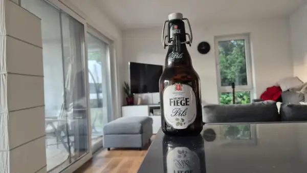 Eine Flasche Moritz Fiege Pils steht auf einem Tisch. Im Hintergrund ein Wohnzimmer mit Sofa und Fernseher.