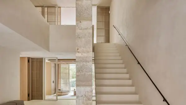 Escaleras de cemento blancas y minimalistas ascienden junto a un pilar de piedra texturizada, conectando niveles en un interior luminoso.