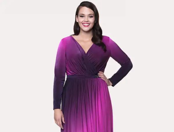 Uśmiechnięta modelka w butik na plus sukienki. Długa, fioletowo-różowa sukienka z dekoltem kopertowym i długimi rękawami.