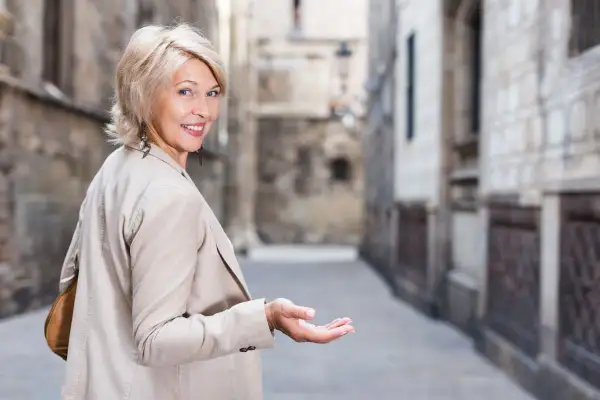 Uśmiechnięta kobieta z blond włosami w modnych, odmładzających krótkich fryzurach bob, zaprasza gestem ręki do wąskiej uliczki.