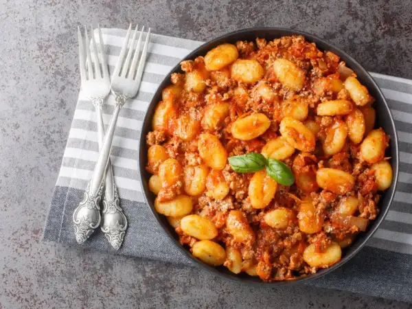 Un plat de gnocchi maison savoureux, nappés d'une sauce bolognaise riche et garnis d'une feuille de basilic frais. Deux fourchettes argentées sont posées sur une serviette à côté.