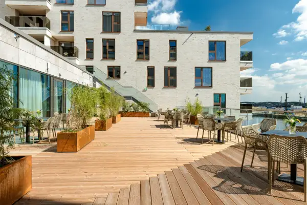 Holzterrasse mit Tischen, Stühlen und Pflanzen vor einem modernen Gebäude mit Blick auf den Hafen.