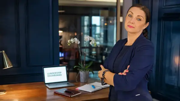 Kobieta w garniturze, z laptopem z napisem "LEADERSHIP DASHBOARD", uczy jak zostać managerem.
