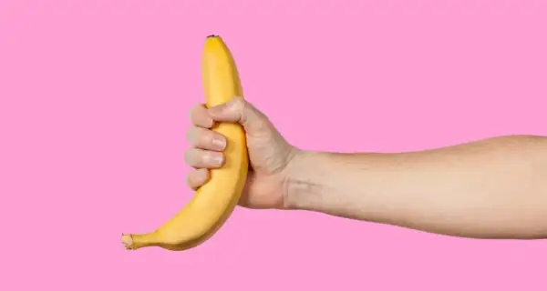 Dłoń trzyma banana na różowym tle. Facet robi sobie dobrze, używając owocu.