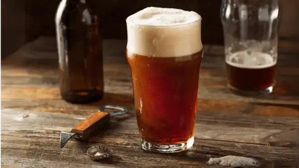 Un bicchiere di birra rossa con schiuma abbondante, accanto a una bottiglia e un apribottiglie. Perfetta per gustare i suoi gradi.