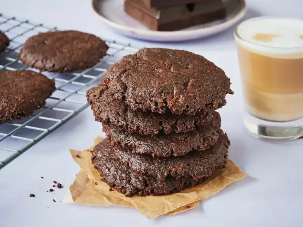 Ein Stapel saftiger brownie cookies, daneben ein Glas Latte Macchiato und ein Teller mit Schokoladenstücken.