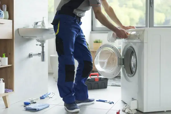 Instalator w niebieskich spodniach podłącza pralkę w łazience. Narzędzia na podłodze.