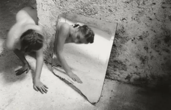 Une femme se contorsionne près d'un miroir brisé, son reflet fragmenté. Une œuvre de Francesca Woodman.