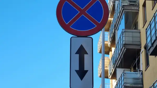 Znak zakaz postoju z tabliczką wskazującą, że zakaz obowiązuje na obu kierunkach. Budynek z balkonami w tle.