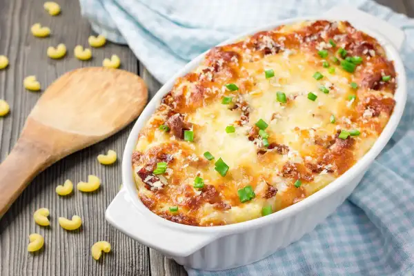 Piatto invitante di pasta al forno alla zucca, dorata e filante, guarnita con erba cipollina. Maccheroni sparsi sul tavolo di legno.