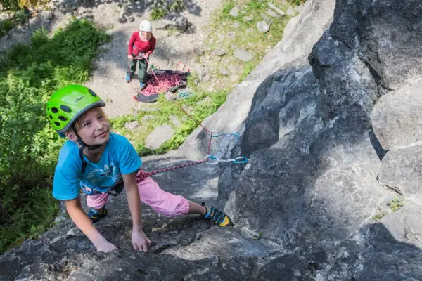 Ein Mädchen mit Helm klettert an einem Felsen, während eine andere Person sichert. Ein tolles Erlebnis beim **Klettern mit Kindern**.
