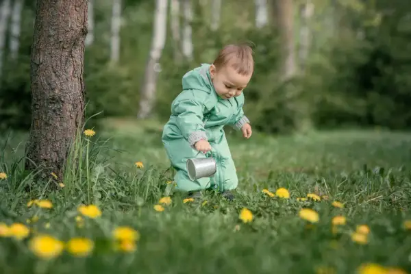 Dziecko w zielonym kombinezonie, z konewką w ręku, odkrywa świat wśród traw i mleczy. To jego **3 skok rozwojowy**, pełen ciekawości i nowych doświadczeń.