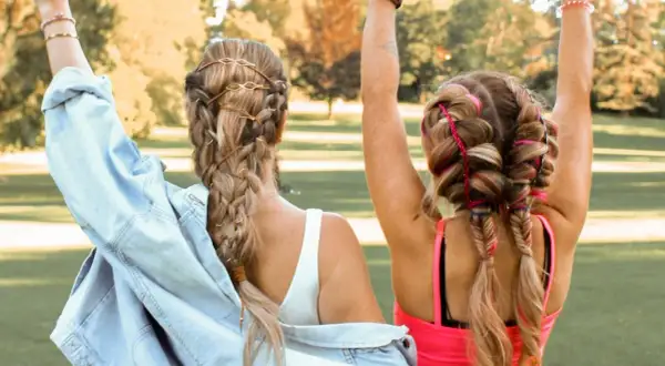 Zwei Mädchen mit geflochtenen Haaren, die sich für festival frisuren einfach eignen. Eine trägt braune Bänder, die andere bunte Fäden.