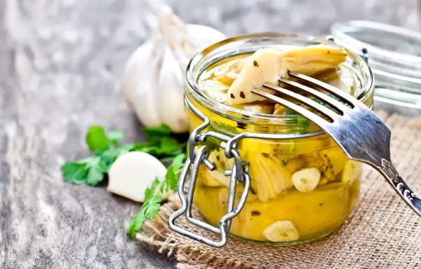 Deliziosi carciofini sott'olio fatti in casa, conservati in un barattolo di vetro con aglio e prezzemolo. Un forchettone ne solleva uno, pronto per essere gustato.