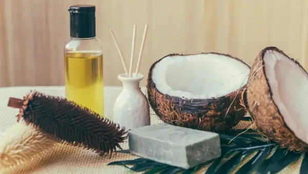 Aceite de coco para el pelo en botella junto a cocos partidos, jabón y cepillo.