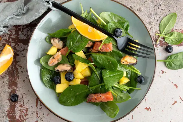Insalata con mango, salmone, cozze e mirtilli, guarnita con spicchio d'arancia e forchetta nera.
