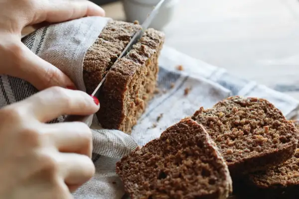 Hände schneiden ein dunkles, gesundes Roggenbrot. Mehrere Scheiben liegen auf einem Leinentuch.