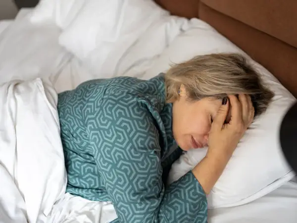 Een vrouw ligt in bed, met haar hand op haar voorhoofd. Ze vraagt zich af: waarom voel ik me zo moe?