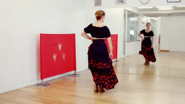 Bailarina con falda flamenca y top negro, practicando una rumbita flamenca frente a un espejo y paneles rojos con detalles dorados.