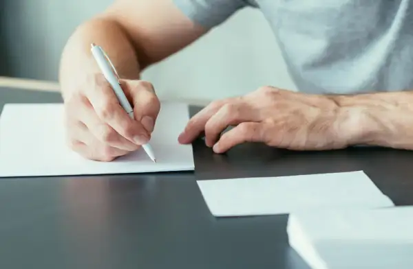 Ręka pisze długopisem na białej kartce, przygotowując się do wysłania listu motywacyjnego.