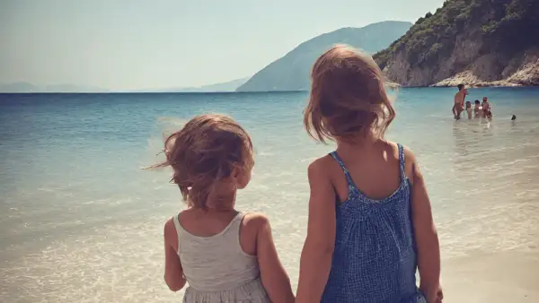 Dwie dziewczynki trzymają się za ręce na plaży, patrząc na morze. W tle widać góry i innych ludzi kąpiących się w wodzie. To idealne wakacje w Albanii z dziećmi.