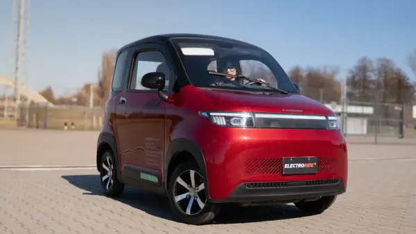 Czerwony, mały samochód elektryczny z kobietą za kierownicą, na tle placu i nieba.