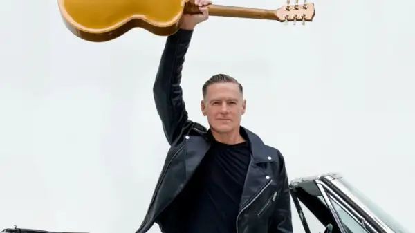 Bryan Adams w skórzanej kurtce unosi gitarę nad głową, gotowy na koncert. Obok klasyczny samochód.